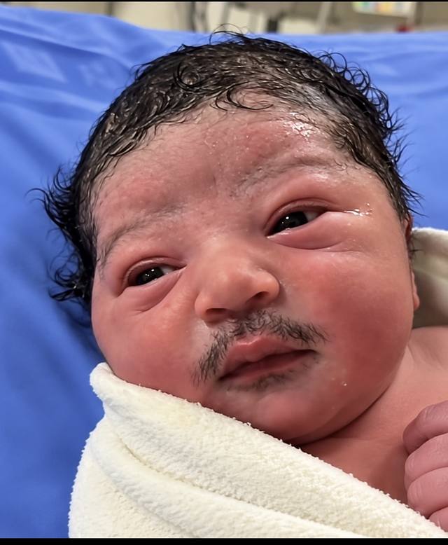 A newborn baby with curly wet hair and dark hair above his upper lip, wrapped in a white towel