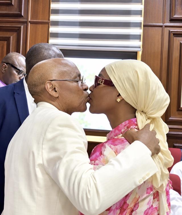 Nasir El-Rufai, in a cream jacket, kisses his wife, who wears a cream headscarf and pink floral dress, in a wood-panelled courtroom
