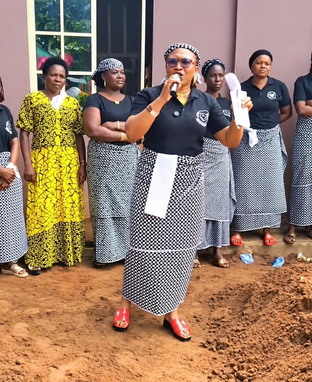 A woman in a black top and checked wrapper speaks into a microphone at a funeral, with other women in matching outfits standing behind her