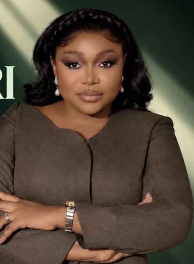Ruth Kadiri in a brown jacket and pearl earrings, arms folded, against a dark green background