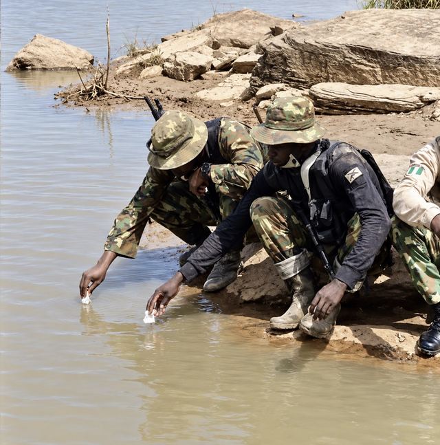 Two Nigerian soldiers in camouflage crouch at a muddy riverbank, dipping pieces of bread into the brown water