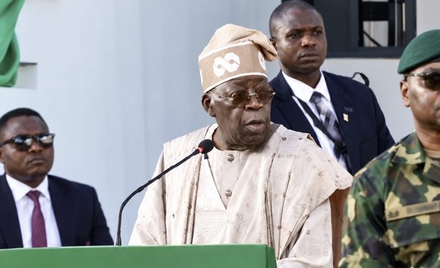 President Bola Tinubu in a cream agbada and cap speaks at a green lectern, flanked by aides and a soldier